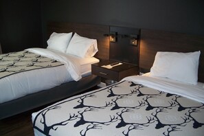 A hotel room with two beds, each with a patterned bedspread, a nightstand with a remote control, and wall-mounted lighting. - Terra Nova Cabins (West Yellowstone)