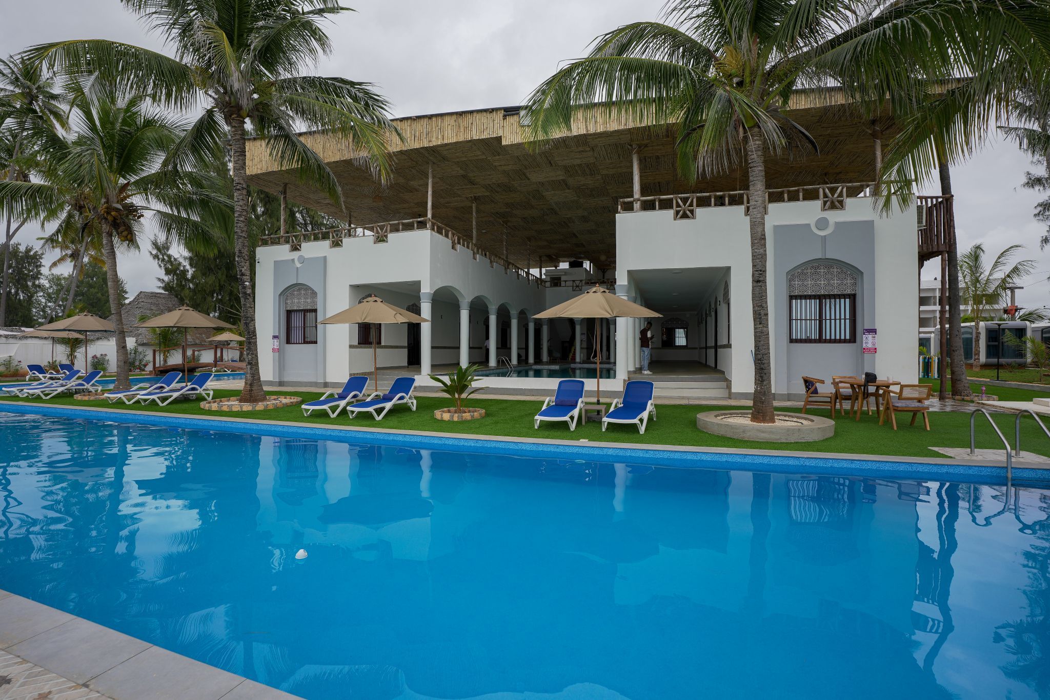Piscina con sillas de descanso y sombrillas, rodeada de palmeras y un edificio con un patio techado.