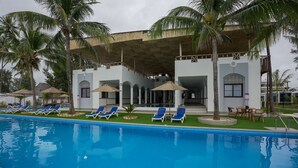 Piscina con sillas de descanso y sombrillas, rodeada de palmeras y un edificio con un patio techado.