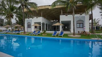 Piscina con sillas de descanso y sombrillas, rodeada de palmeras y un edificio con un patio techado.