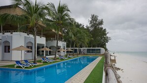 Un área de piscina con sillas de descanso y sombrillas, una playa al fondo, y un edificio con techo de paja.