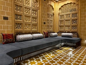 A room with a sofa set against intricately carved walls, a patterned floor, and a power outlet on the wall. - Gulaab Haveli (Jaisalmer)