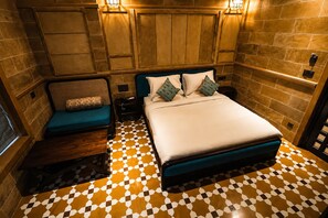 A bedroom with a checkered floor, a bed with pillows, a nightstand, and a seating area with a cushion. - Gulaab Haveli (Jaisalmer)