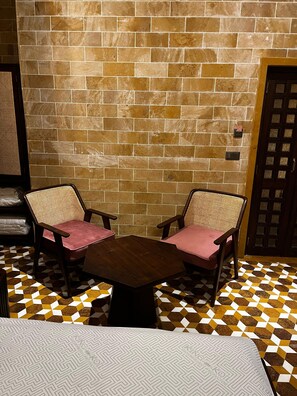 A room with a brick wall, two wooden chairs with pink cushions, a wooden table, and a patterned floor. - Gulaab Haveli (Jaisalmer)