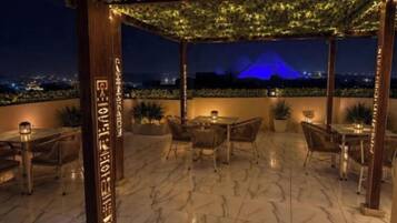 An outdoor seating area with tables and chairs, a pergola with string lights, and a view of a pyramid at night.