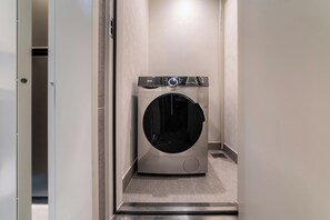 A compact, stainless steel front-loading washing machine in a small, enclosed laundry room with a tiled floor.