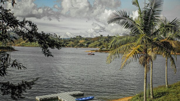 A calm lake with a boat, a dock with a kayak, and palm trees. - La Pausa Hotelbistro (San Luis)