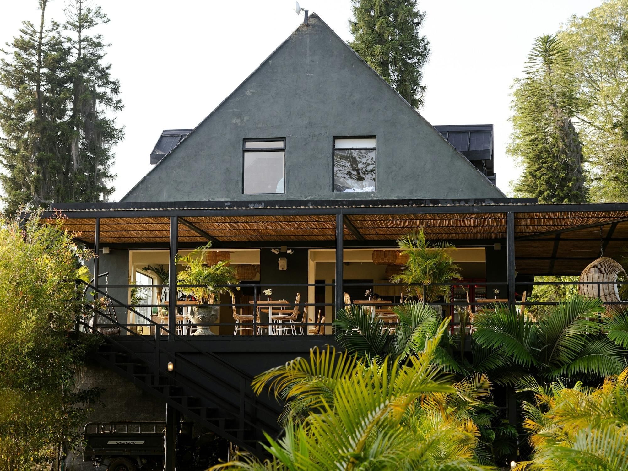 Un edificio moderno con techo triangular, grandes ventanales y un área de estar exterior cubierta con mesas y sillas.