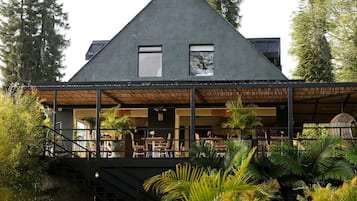 Un edificio moderno con techo triangular, grandes ventanales y un área de estar exterior cubierta con mesas y sillas.