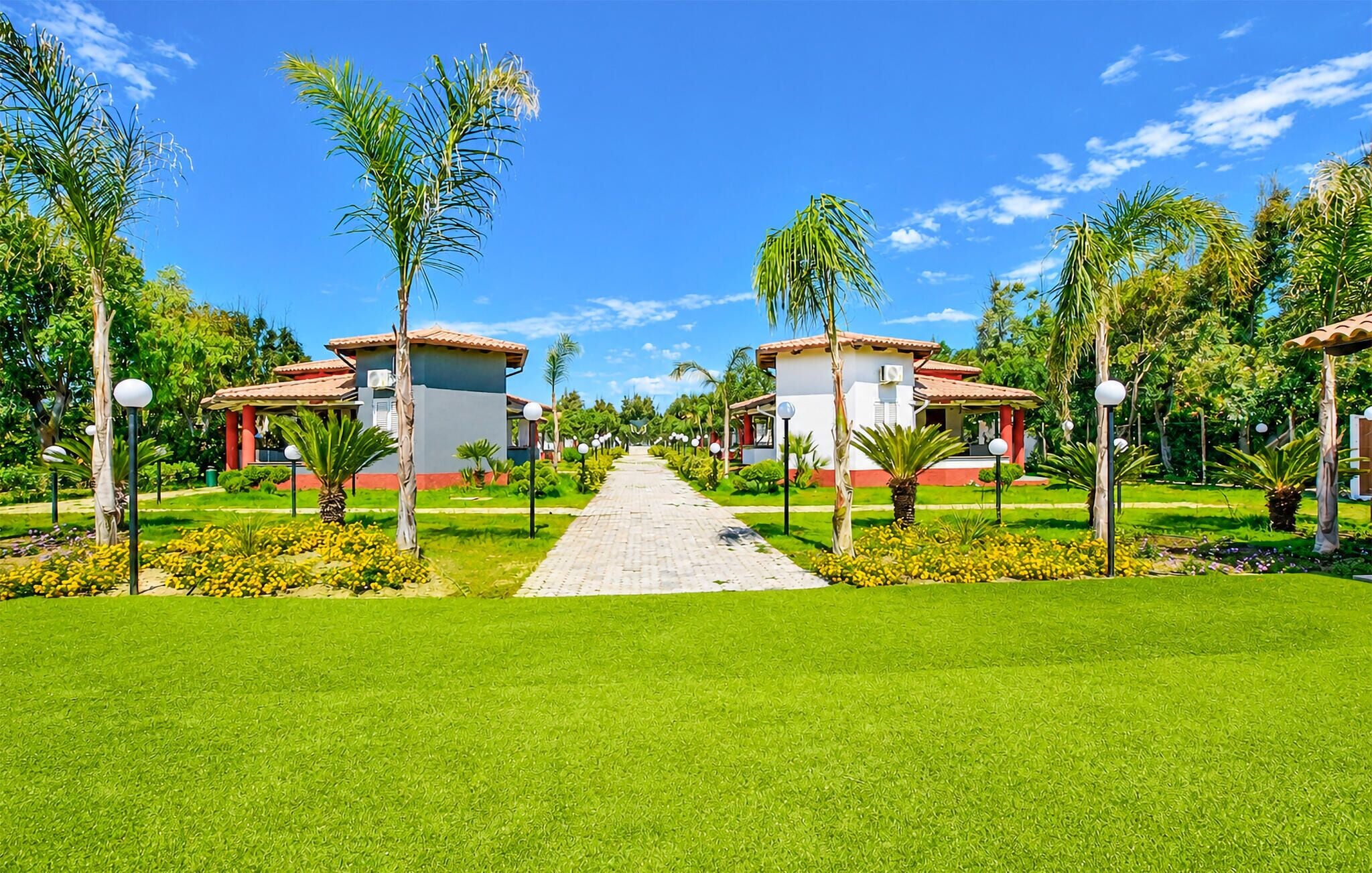 A pathway leading to a building with a red roof, surrounded by greenery and palm trees.