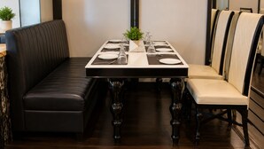A dining area with a rectangular table set for four, flanked by black leather booths and beige upholstered chairs. - The Regaal - 5 mins from Railway Station (Jalgaon)