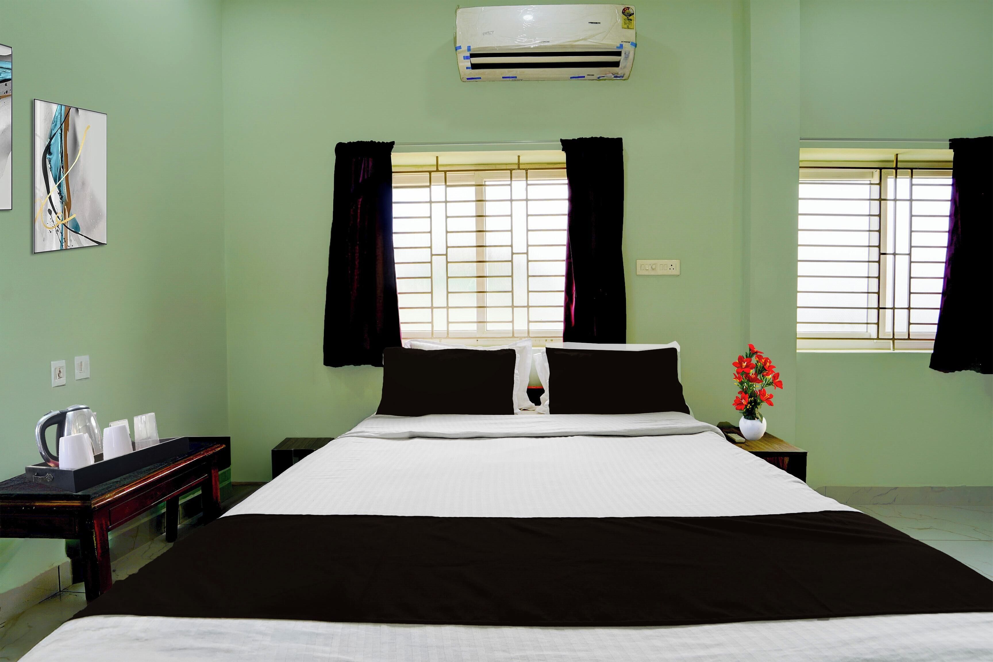 A hotel room with a bed, a bedside table with a kettle and cups, a nightstand, and a window with curtains.