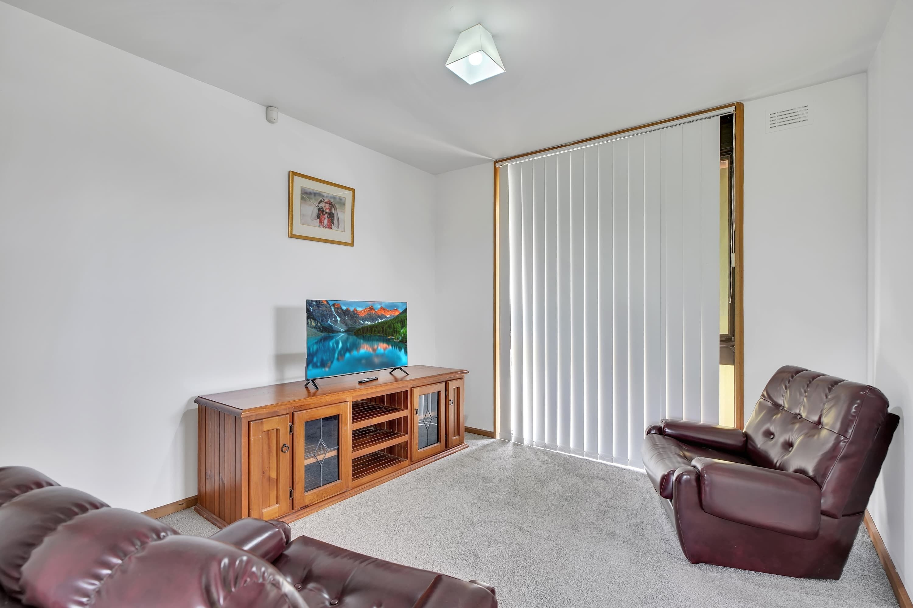 A living room with a television on a wooden cabinet, a brown leather sofa, and a large window with vertical blinds.