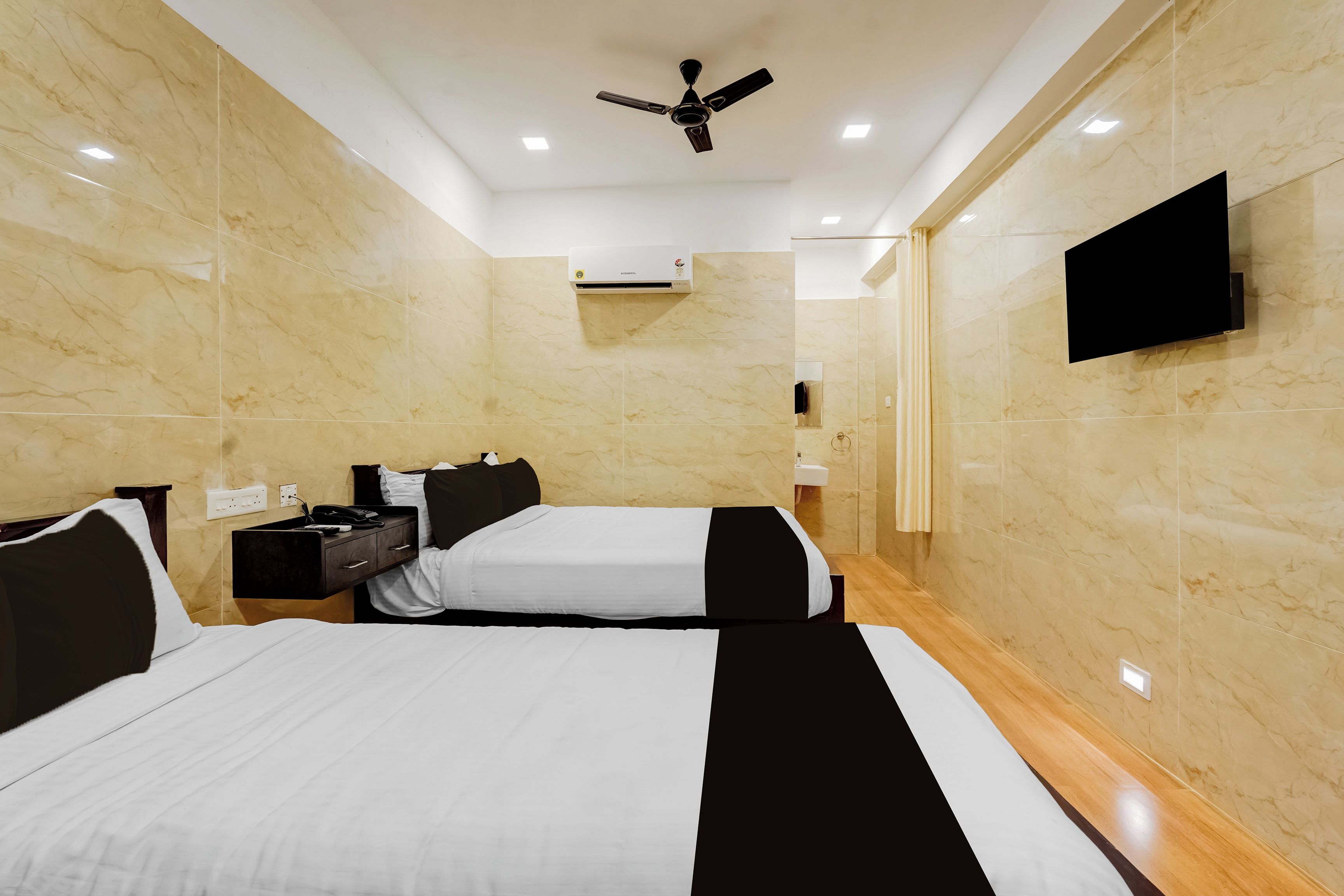 A hotel room with two beds, a TV, a ceiling fan, and a bathroom with a sink and mirror.