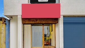 Una entrada de edificio con toldo rojo, persiana azul cerrada y un interior visible con una silla y una mesa.