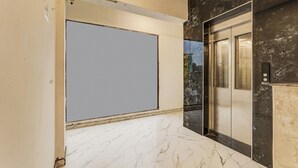 Un pasillo con piso de mármol, una gran pantalla proyectora vacía y un ascensor con acabado metálico.
