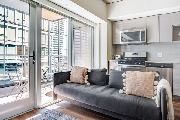 Una sala de estar moderna con un sofá gris, una cocina con electrodomésticos de acero inoxidable y un amplio ventanal que da a una zona de estar exterior.
