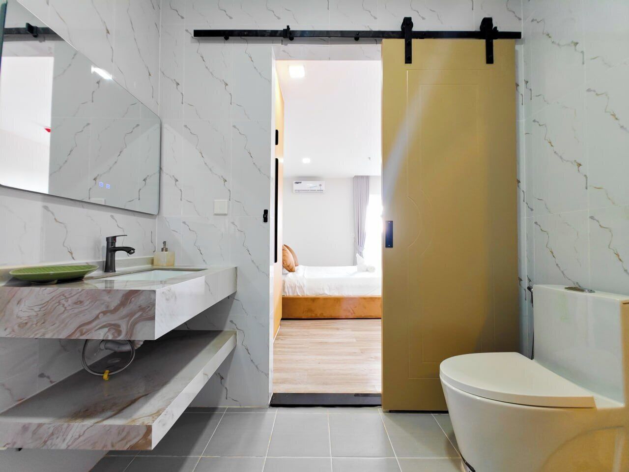 A modern bathroom with a marble vanity, a toilet, and a bed in the adjacent room.