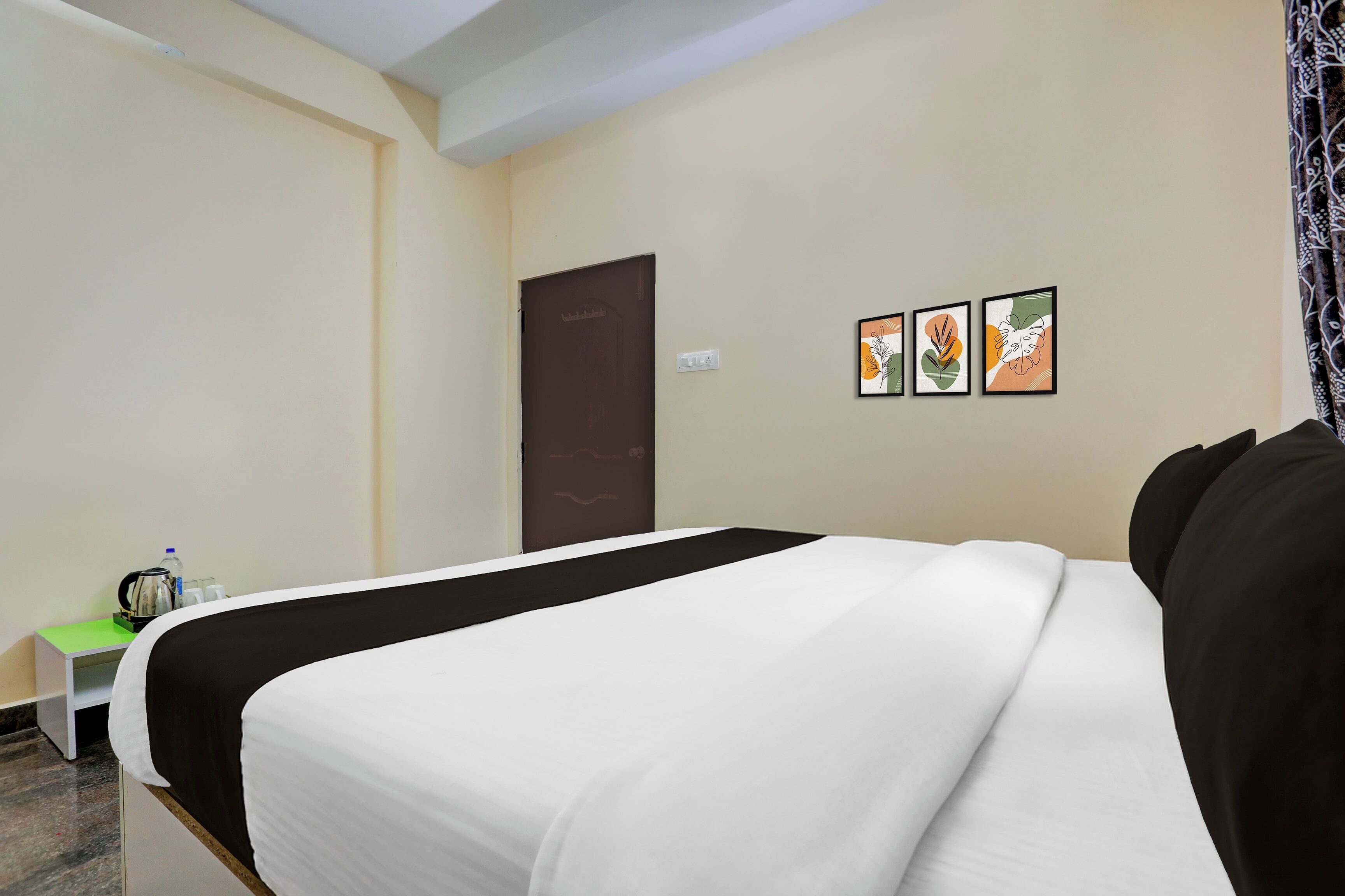Habitación de hotel con una cama, una mesita con hervidor y tazas, y una puerta. Hay tres cuadros enmarcados en la pared.