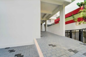 A corridor with a tiled floor, a white wall, and a red and green building in the background. - Collection O Valiyavila Junction (Thiruvananthapuram)