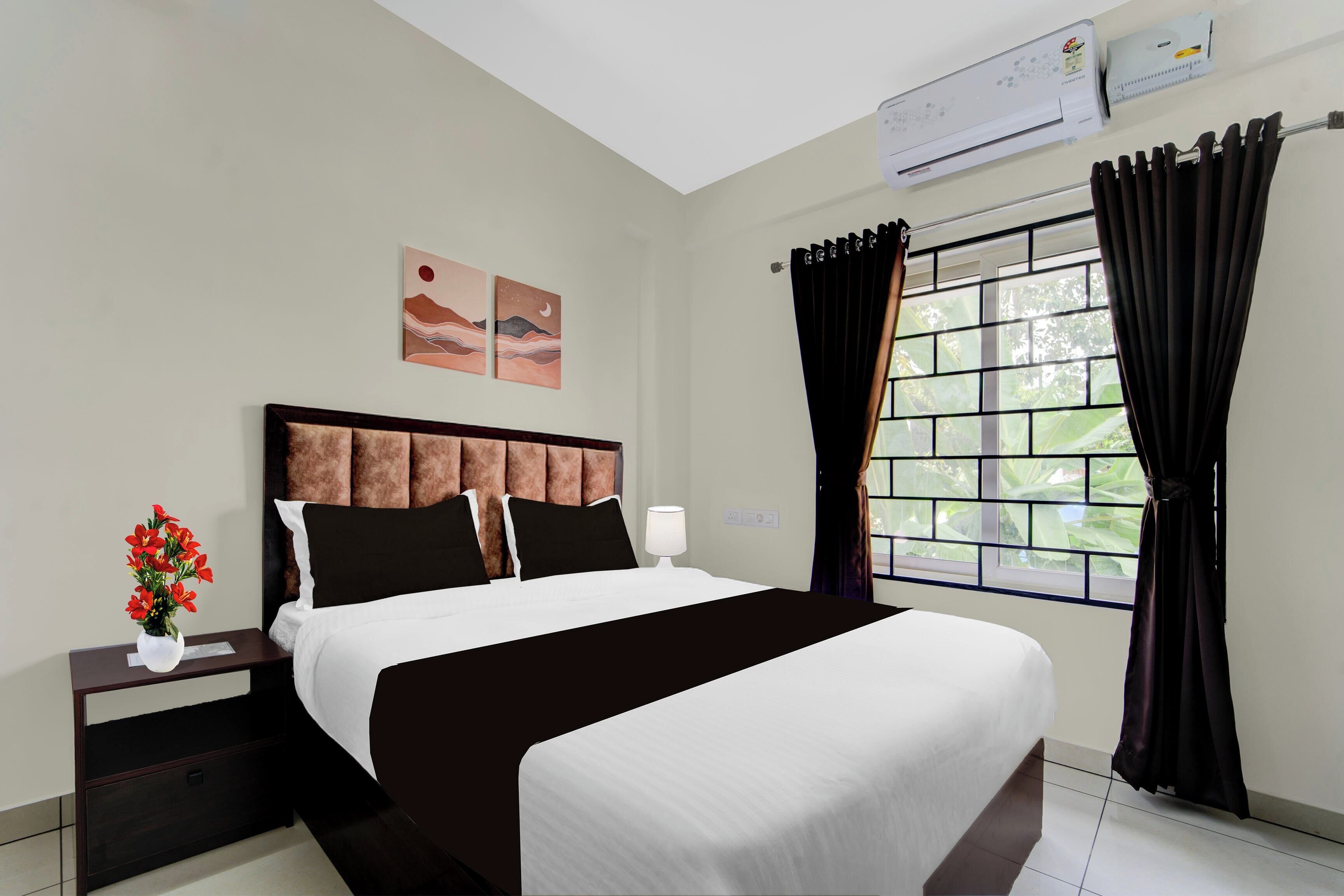 A hotel room with a large bed, a nightstand, a vase of flowers, a window with curtains, and an air conditioning unit.