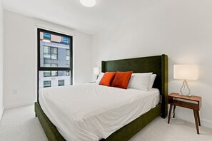 Ein Hotelzimmer mit einem großen Bett, einem Nachttisch mit Lampe und einem Fenster mit Blick auf Gebäude.