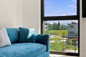 A modern living room with a large window, a blue sofa, and a view of a building and greenery outside. - Stairway to Heaven - 3 Bed Condo - Rooftop Hot Tub (Revelstoke)