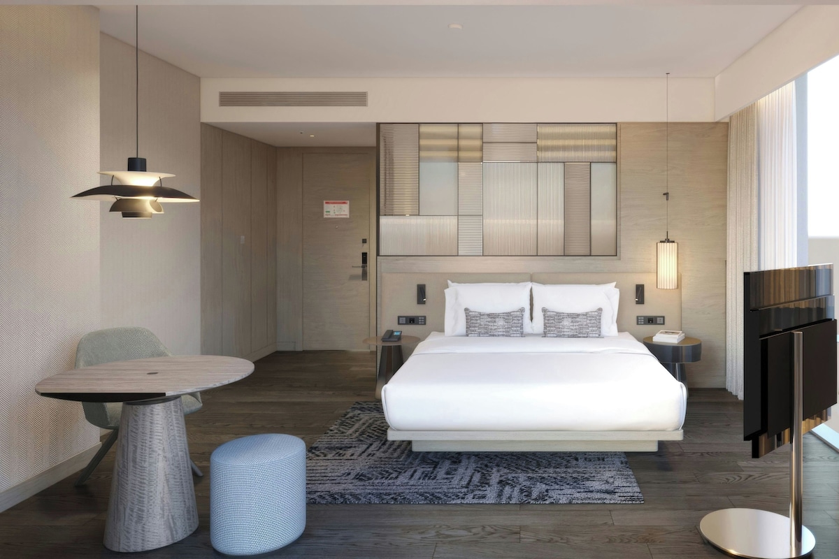 A modern hotel room with a large bed, a desk, a chair, a TV, and a large window.