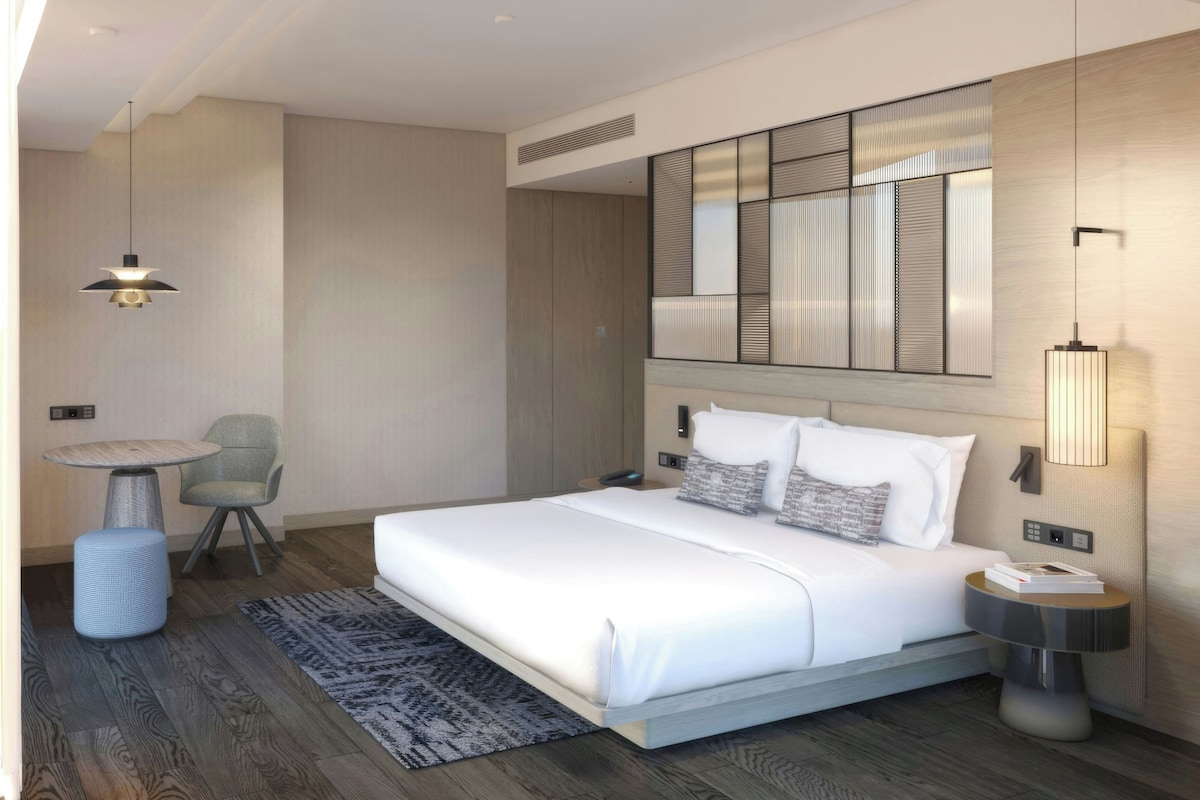 A modern hotel room with a large bed, a small round table, a chair, a bedside lamp, and a wall-mounted control panel.