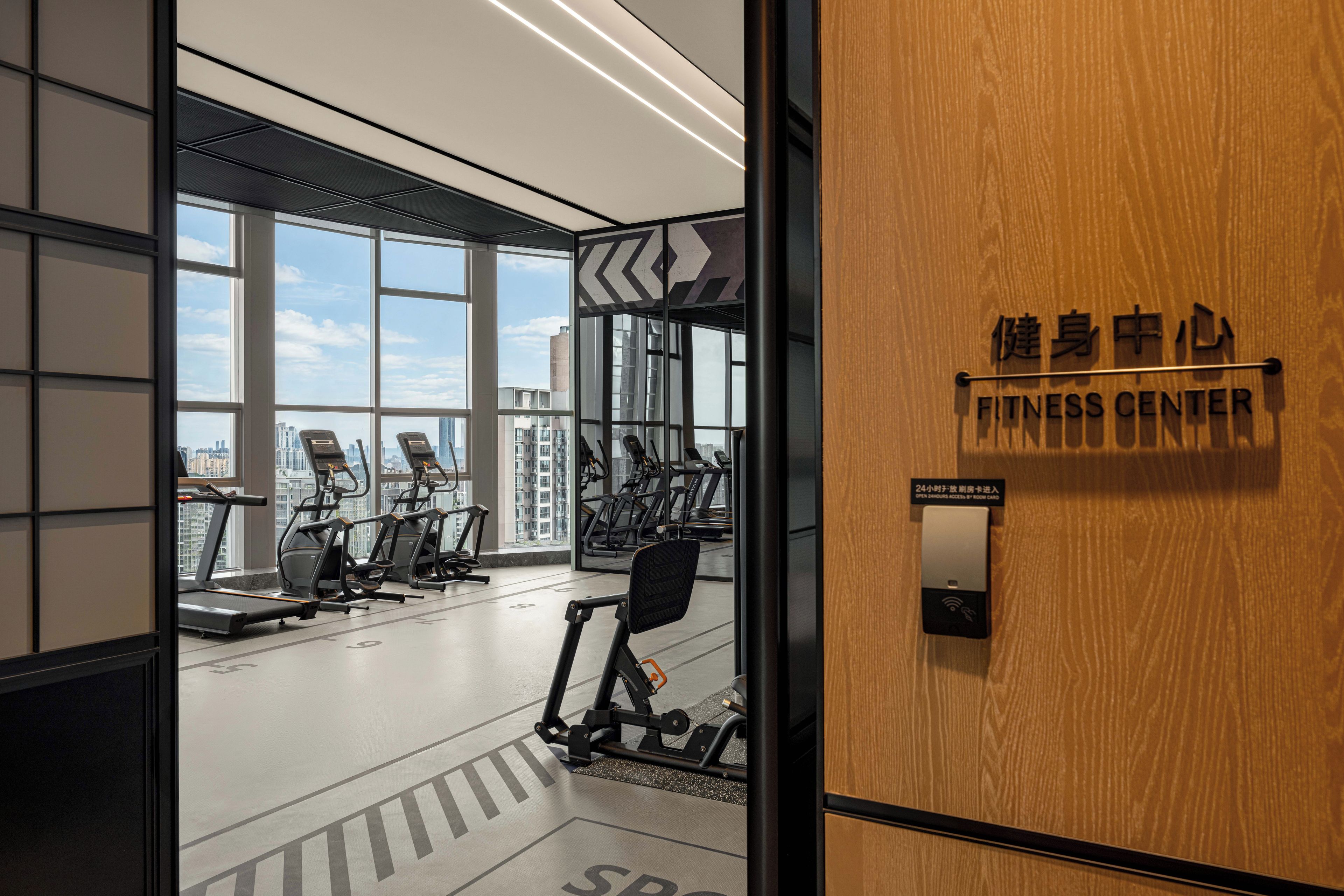 Une salle de sport équipée de matériel d’exercice, dotée de grandes fenêtres et d’une enseigne indiquant qu’elle est accessible 24 heures sur 24.