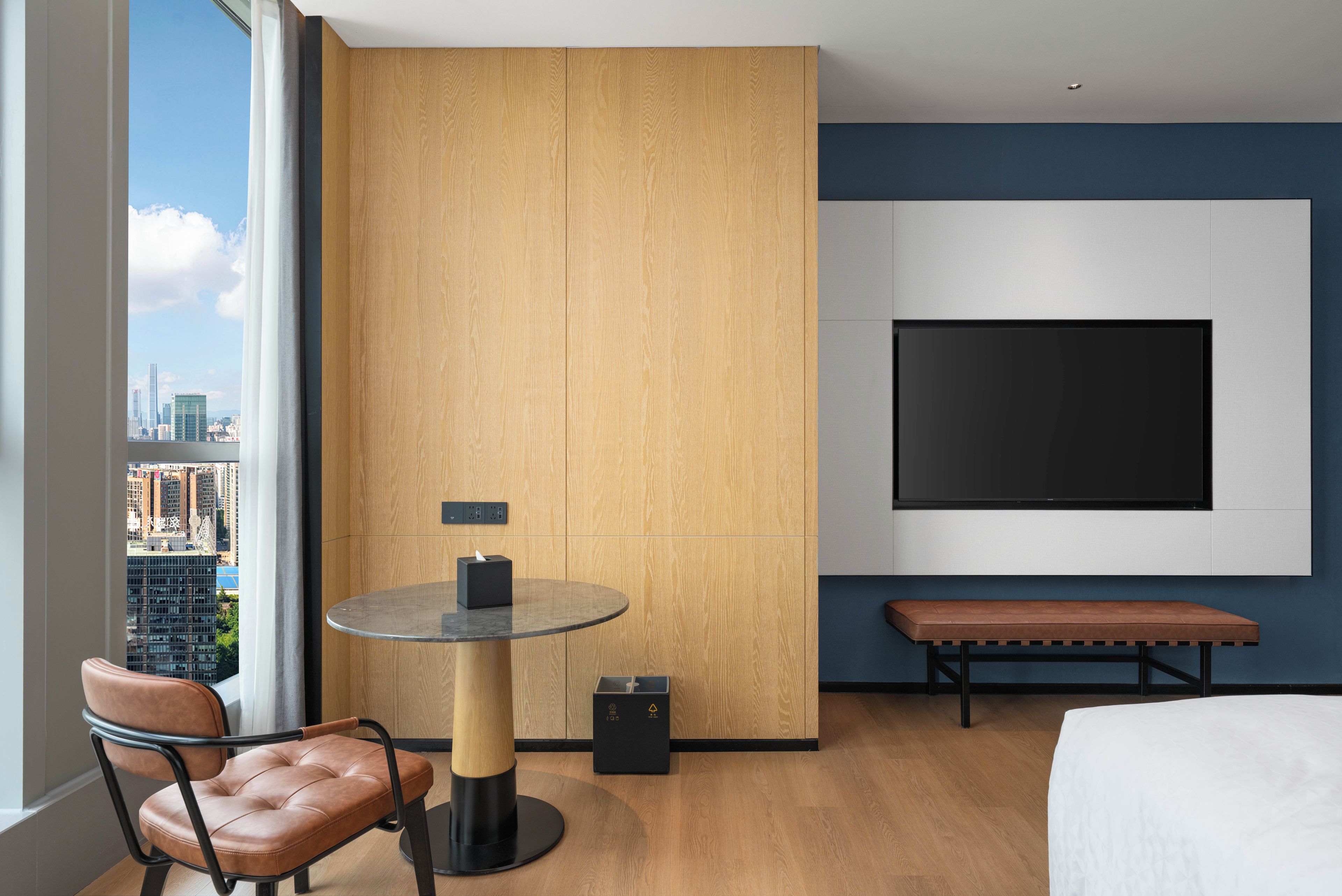 A hotel room with a large window, a round table with a tissue box, a bench, and a flat-screen TV mounted on the wall.