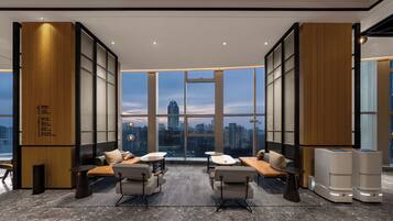 A modern hotel room with large windows, a city view, a seating area with a sofa and chairs, and a small table.