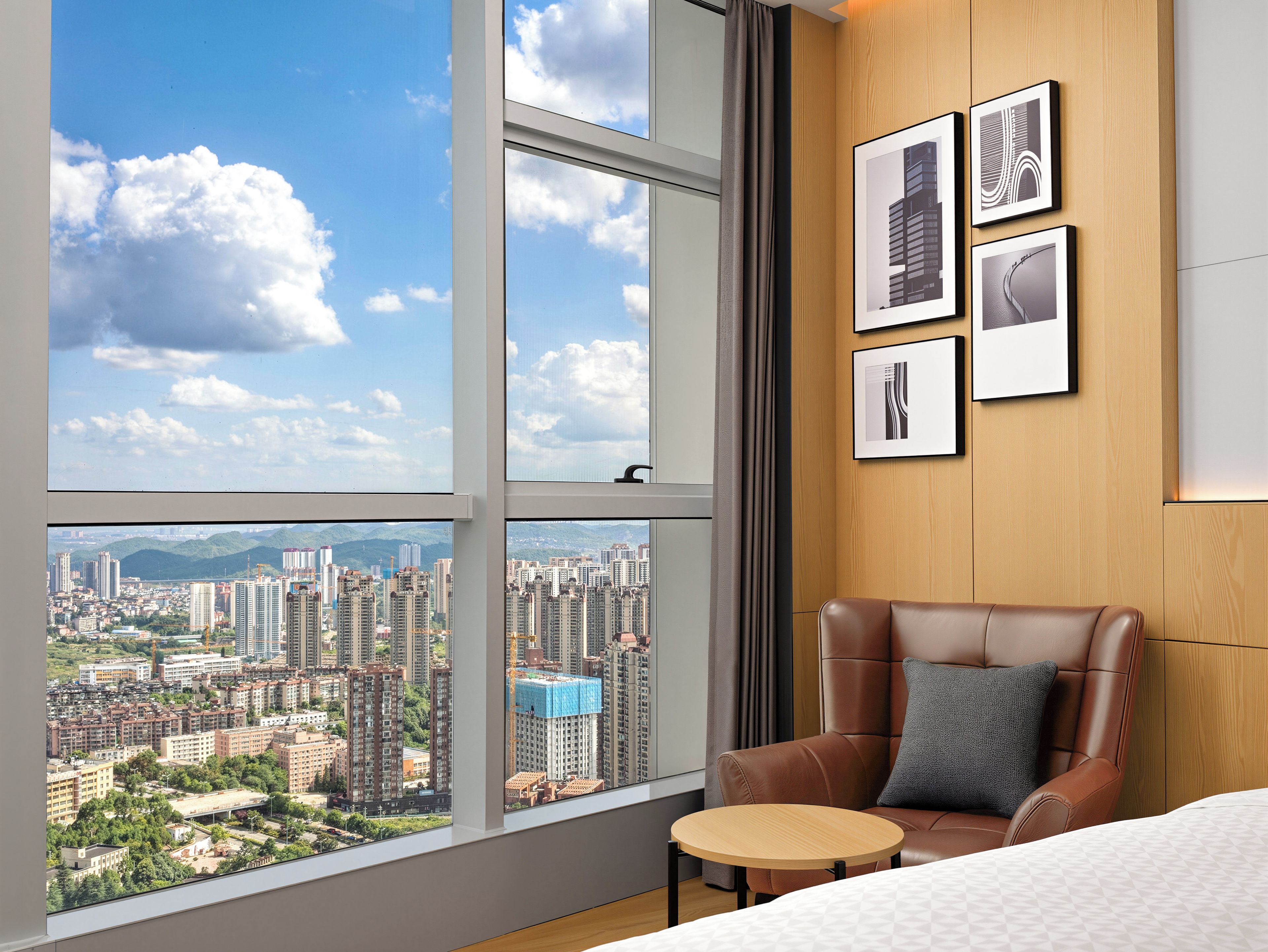 A hotel room with a large window, a brown leather chair, a small round table, and framed pictures on the wall.