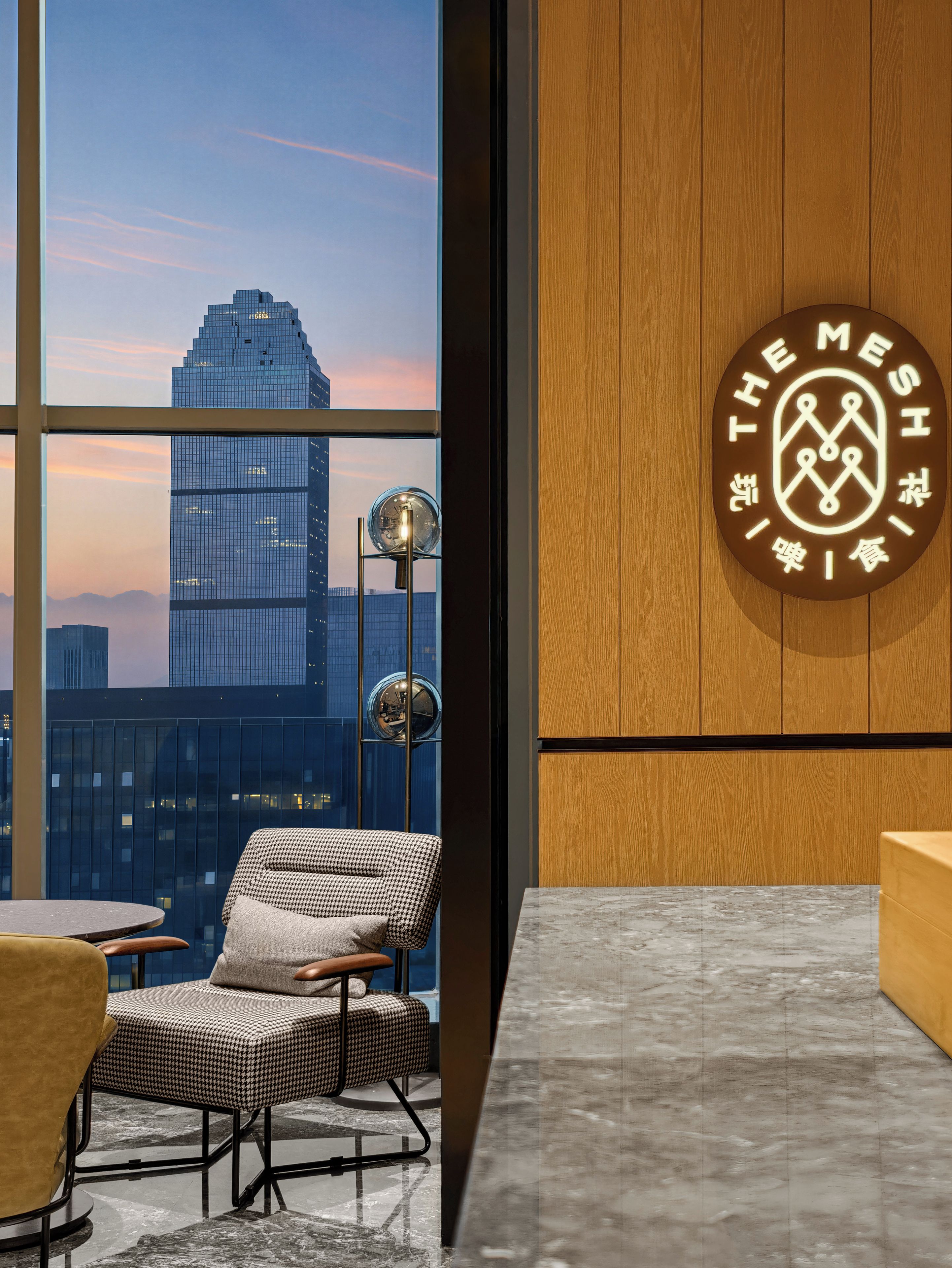 A modern interior with a view of a cityscape through large windows, a houndstooth patterned chair, and a wooden wall with a logo.