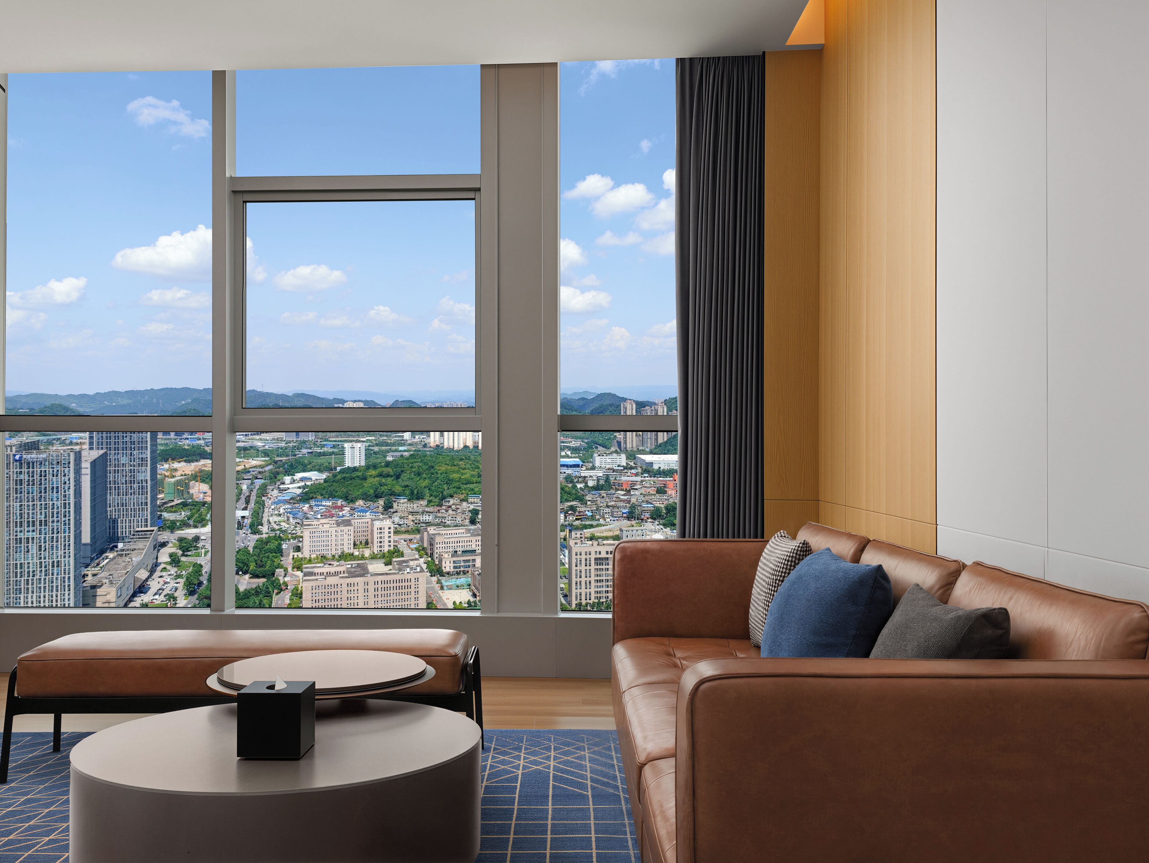 A modern living room with a leather sofa, a round coffee table, and a view of a cityscape through large windows.