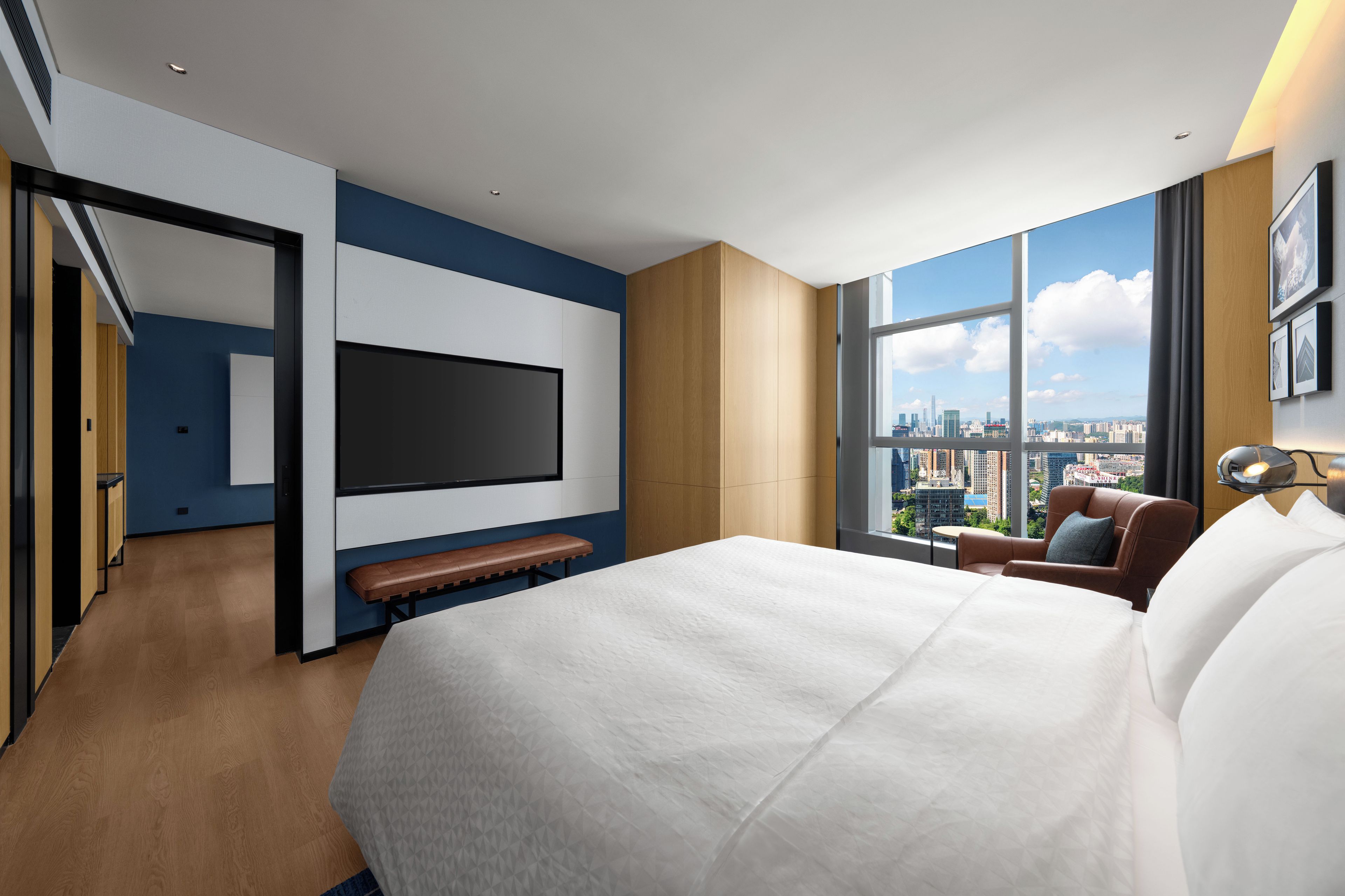 A modern hotel room with a large bed, a flat-screen TV, a seating area with a bench, and a city view through large windows.