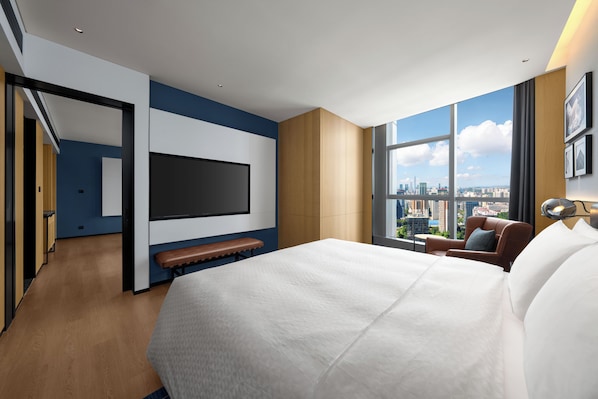 A modern hotel room with a large bed, a flat-screen TV, a seating area with a bench, and a city view through large windows.