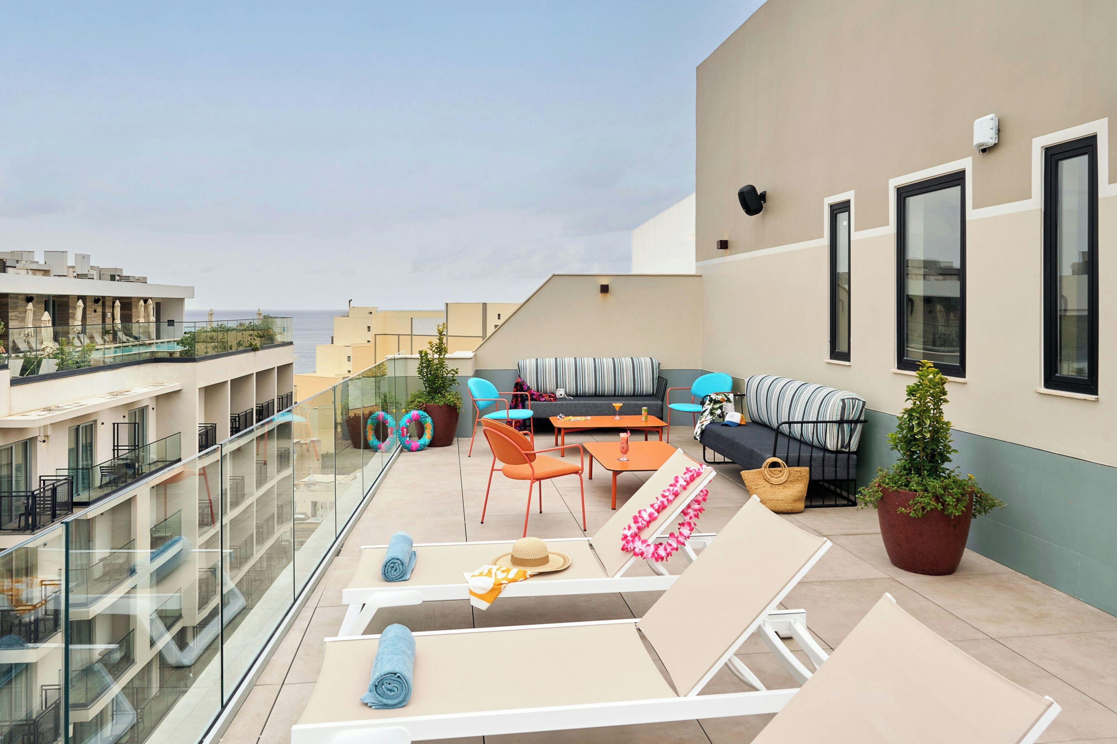 Una terraza en la azotea con barandilla de vidrio, zona de estar exterior con sillones y sofá, plantas en macetas y vista al mar.