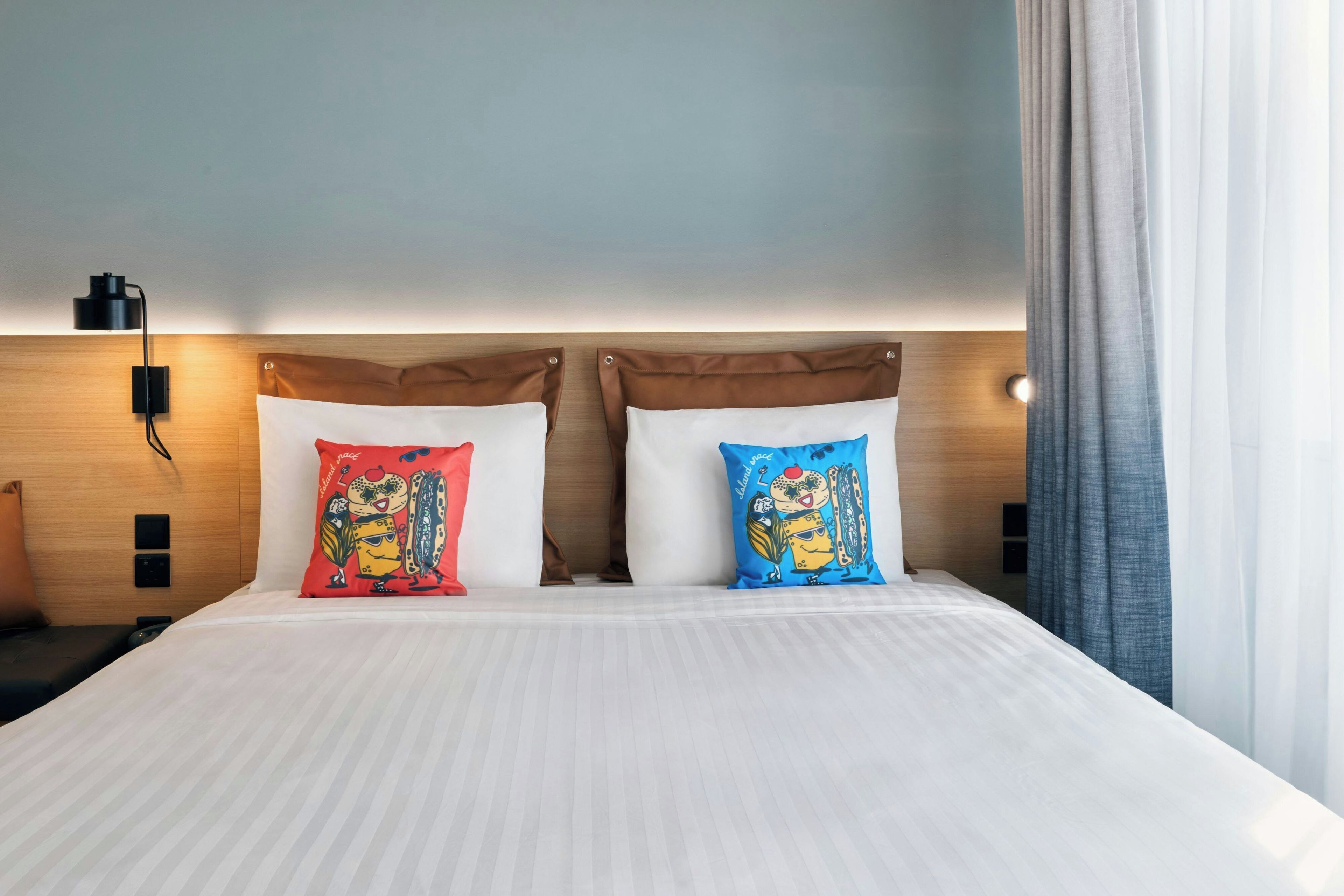 A hotel room with a bed, two pillows with cartoon characters, a nightstand, a lamp, and a window with curtains.
