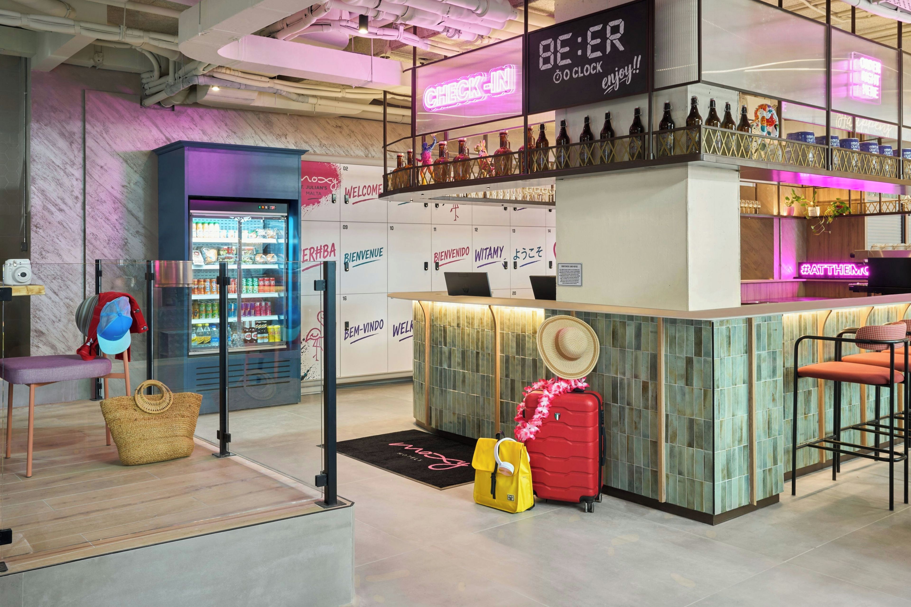 A bar area with a neon sign, a refrigerator with drinks, and a seating area with a hat and luggage.
