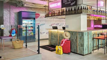 A bar area with a neon sign, a refrigerator with drinks, and a seating area with a hat and luggage.