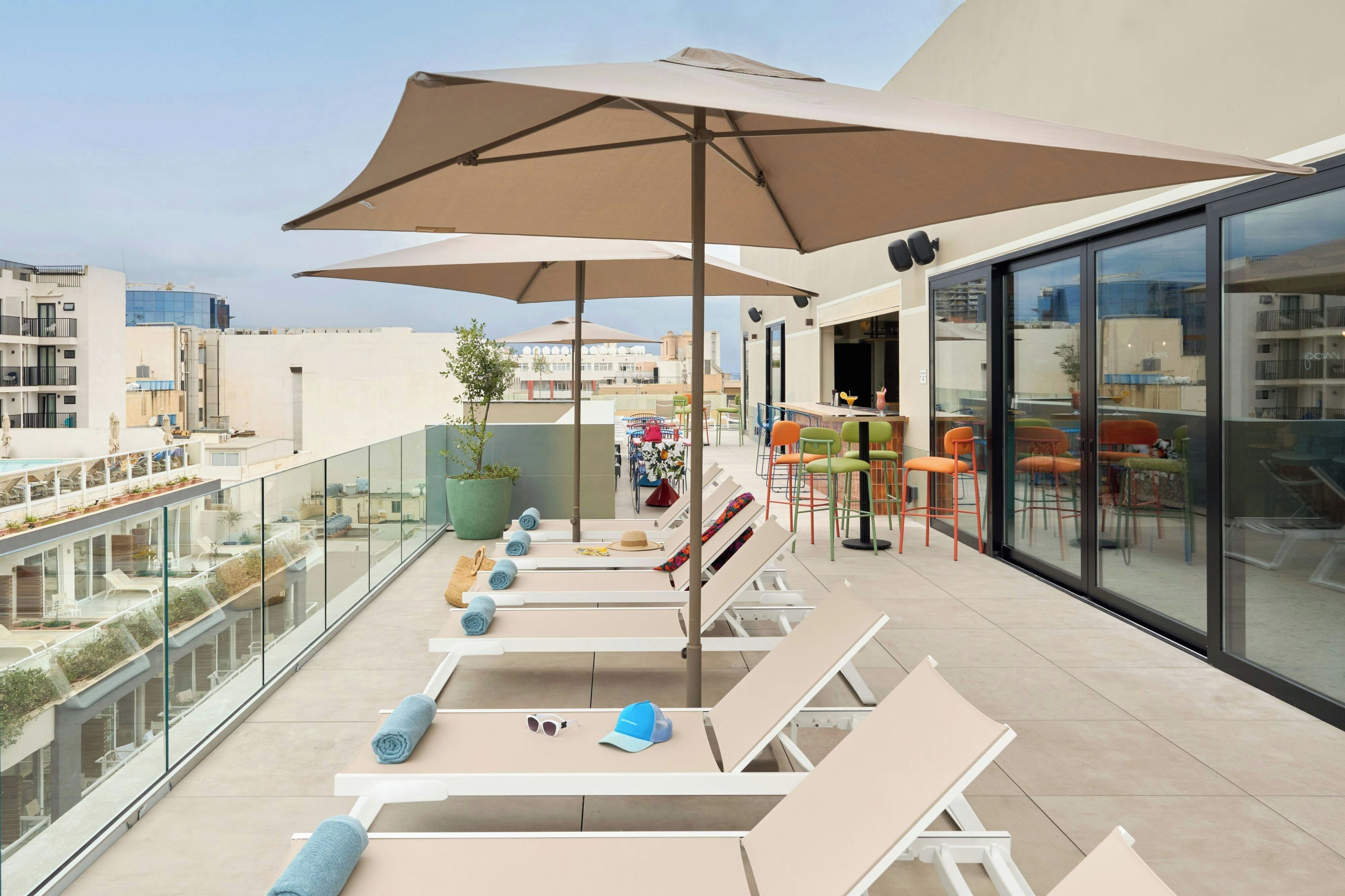 Una terraza en la azotea con sillones de sol, un gran parasol y balaustrada de cristal con vistas a la ciudad.