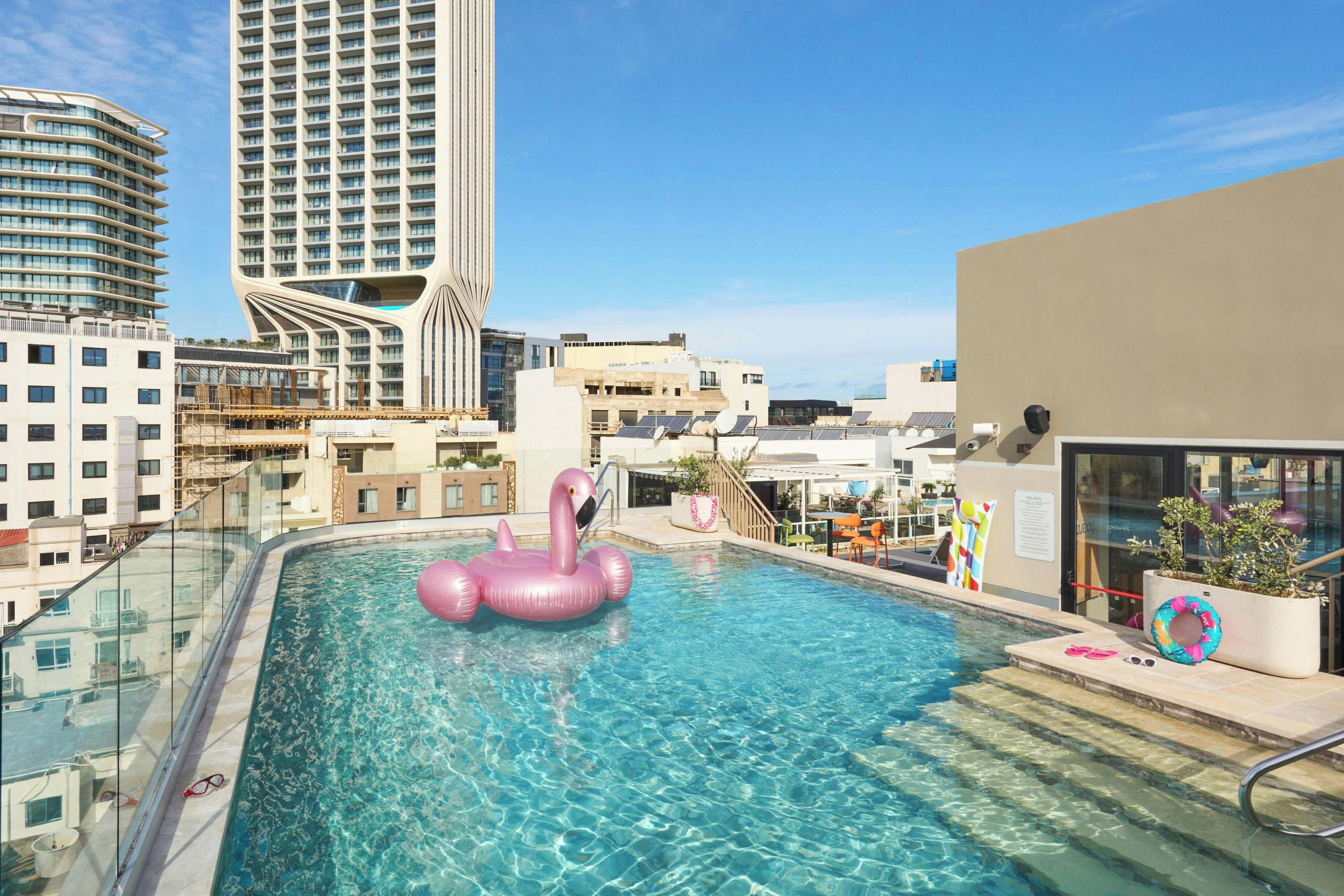 Una piscina en la azotea con un flotador de flamenco rosa, rodeada de edificios modernos y cielos despejados.