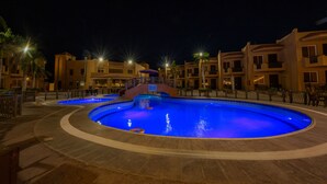 Un área de piscina nocturna con varias piscinas, rodeada de edificios con balcones y mobiliario de exterior.