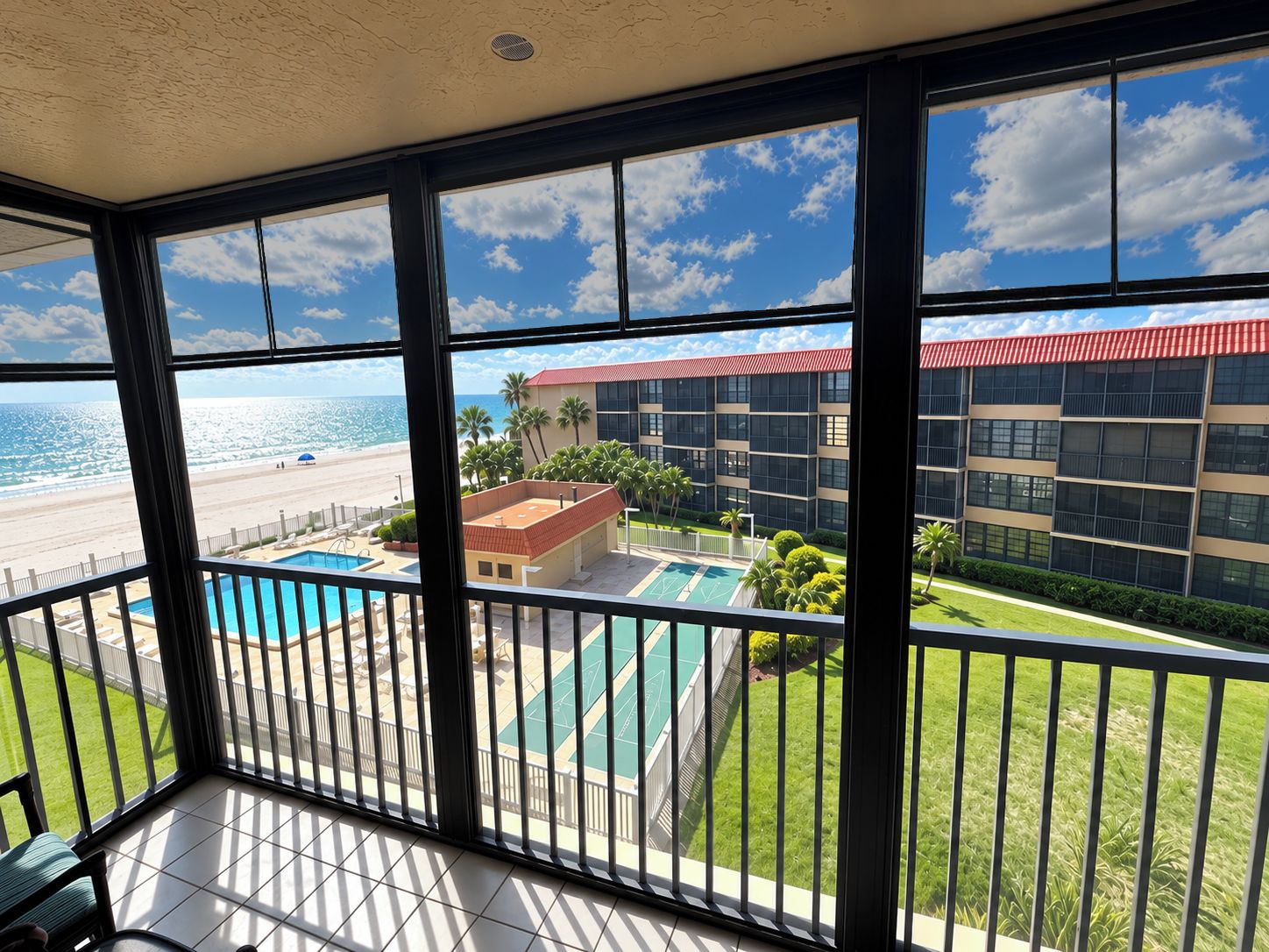 A balcony with a view of a beach, a pool, and a building with a red roof.