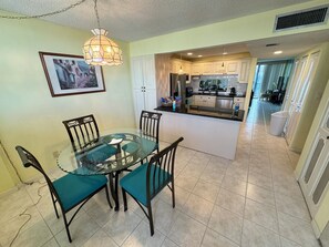 Comedor con una mesa y sillas de vidrio, cocina al fondo y un cuadro en la pared.
