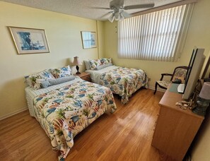 A room with two beds, a ceiling fan, a window with blinds, and a desk with a lamp. - Beachfront Condo (Redington Shores)