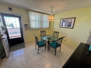 A dining area with a glass table and chairs, a hanging light fixture, a door with a view to the outside, and a framed picture on the wall. - Beachfront Condo in Redington Shores - Available January - April (Redington Shores)