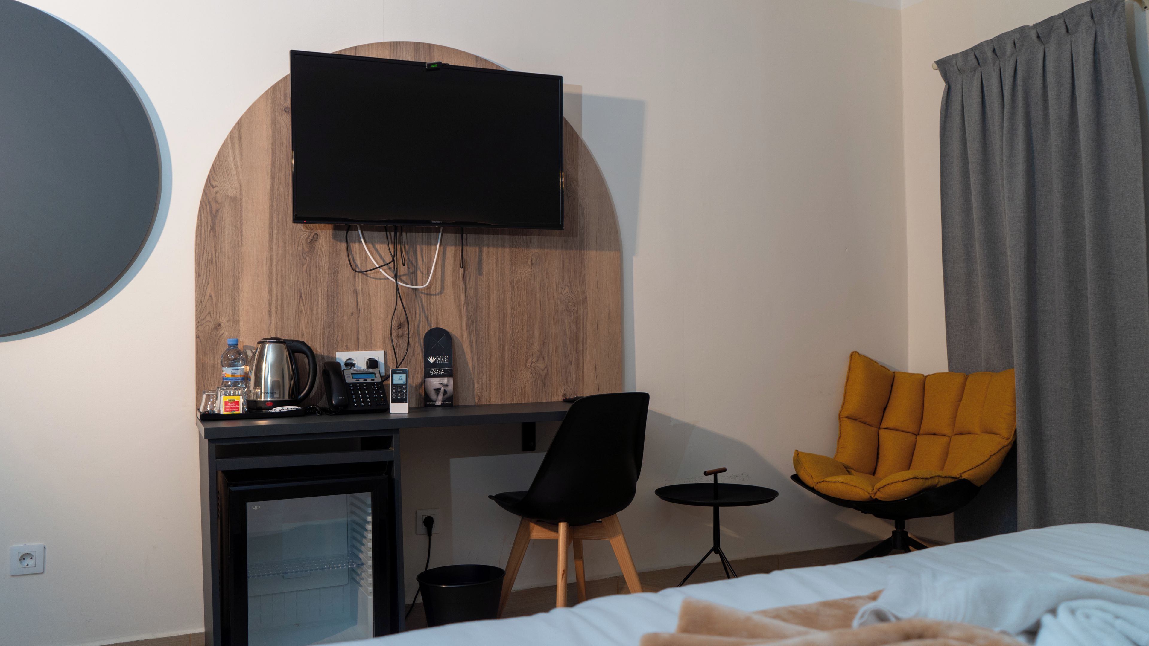 A hotel room with a bed, a desk with a computer and phone, a television mounted on a wooden wall, and a yellow armchair.