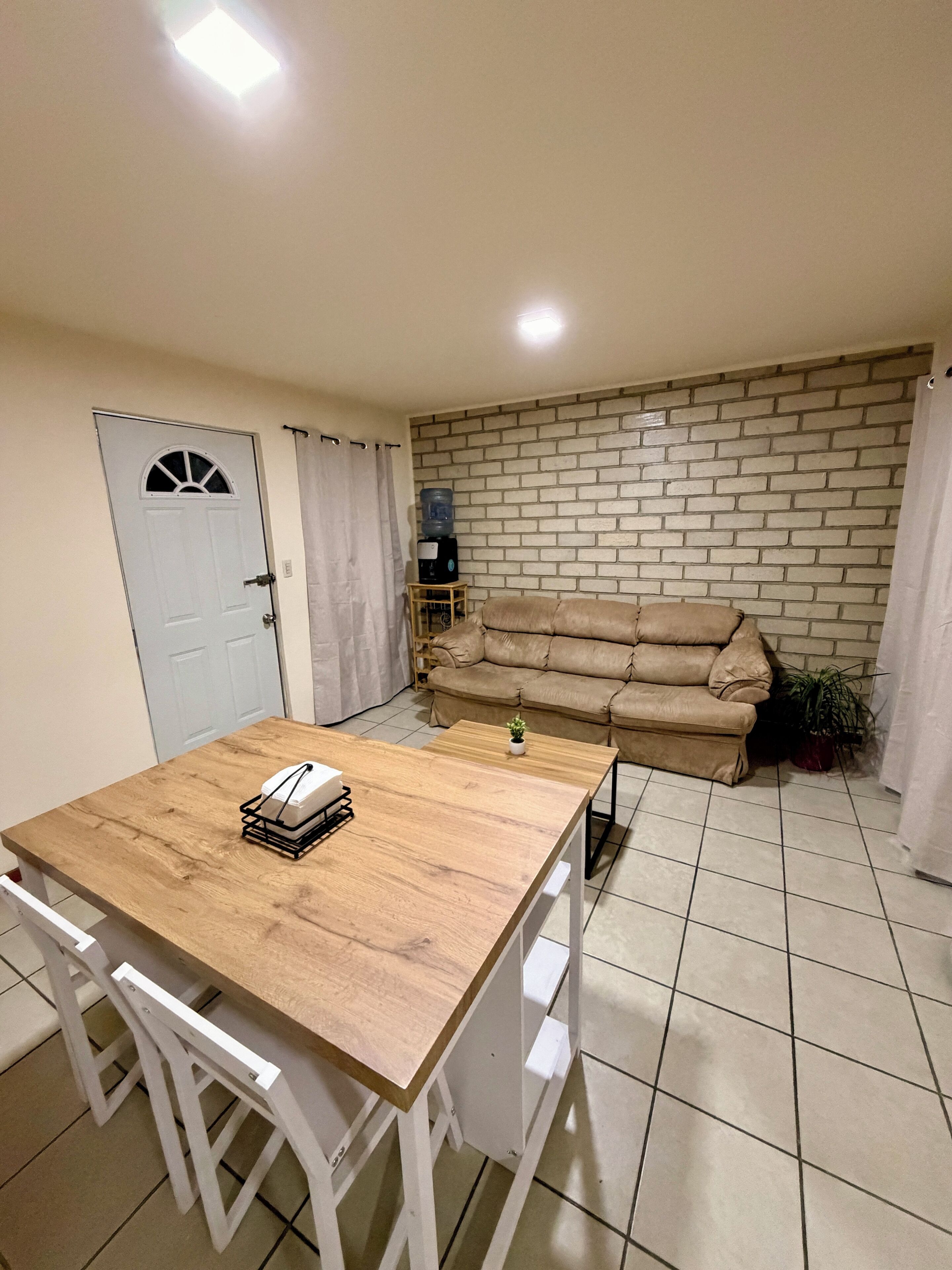 A room with a wooden table, white chairs, a beige sofa, a water dispenser, and a brick-patterned wall.
